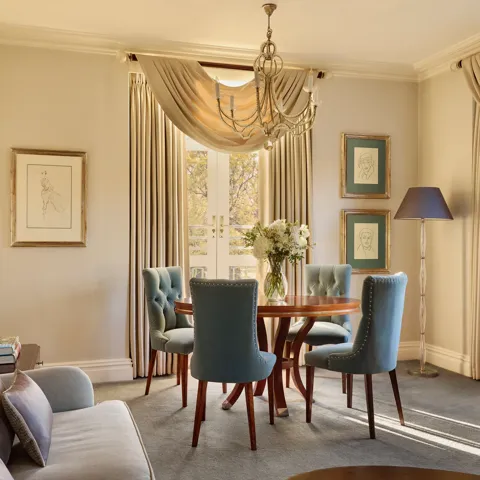 Elegant dining space with a round wooden table surrounded by teal upholstered chairs, crystal chandelier above, and French doors draped with flowing curtains