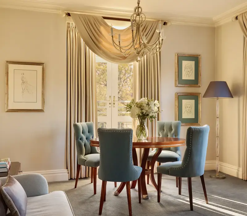 Elegant dining space with a round wooden table surrounded by teal upholstered chairs, crystal chandelier above, and French doors draped with flowing curtains