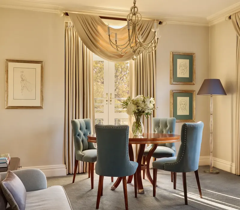Elegant dining space with a round wooden table surrounded by teal upholstered chairs, crystal chandelier above, and French doors draped with flowing curtains