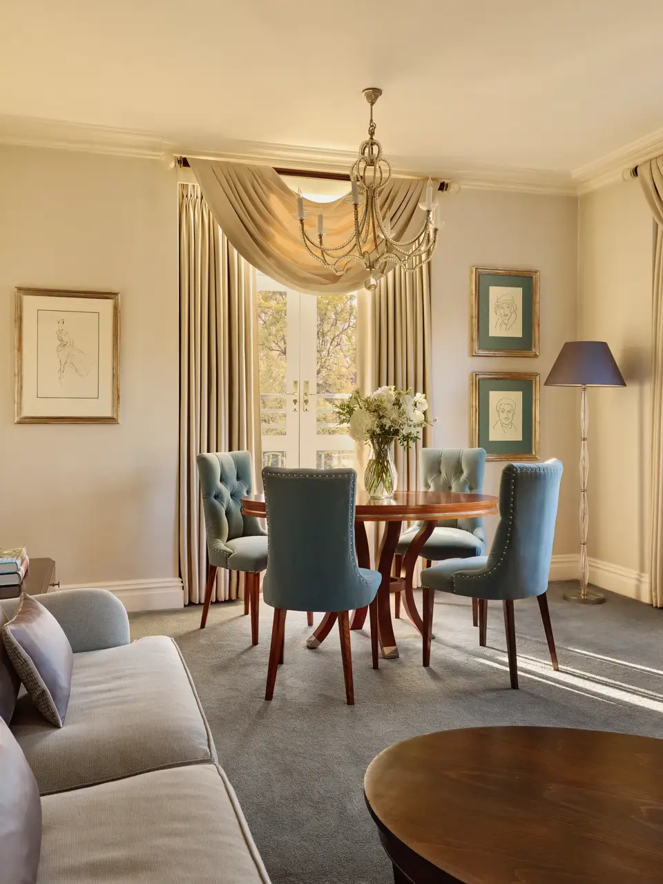 Elegant dining space with a round wooden table surrounded by teal upholstered chairs, crystal chandelier above, and French doors draped with flowing curtains