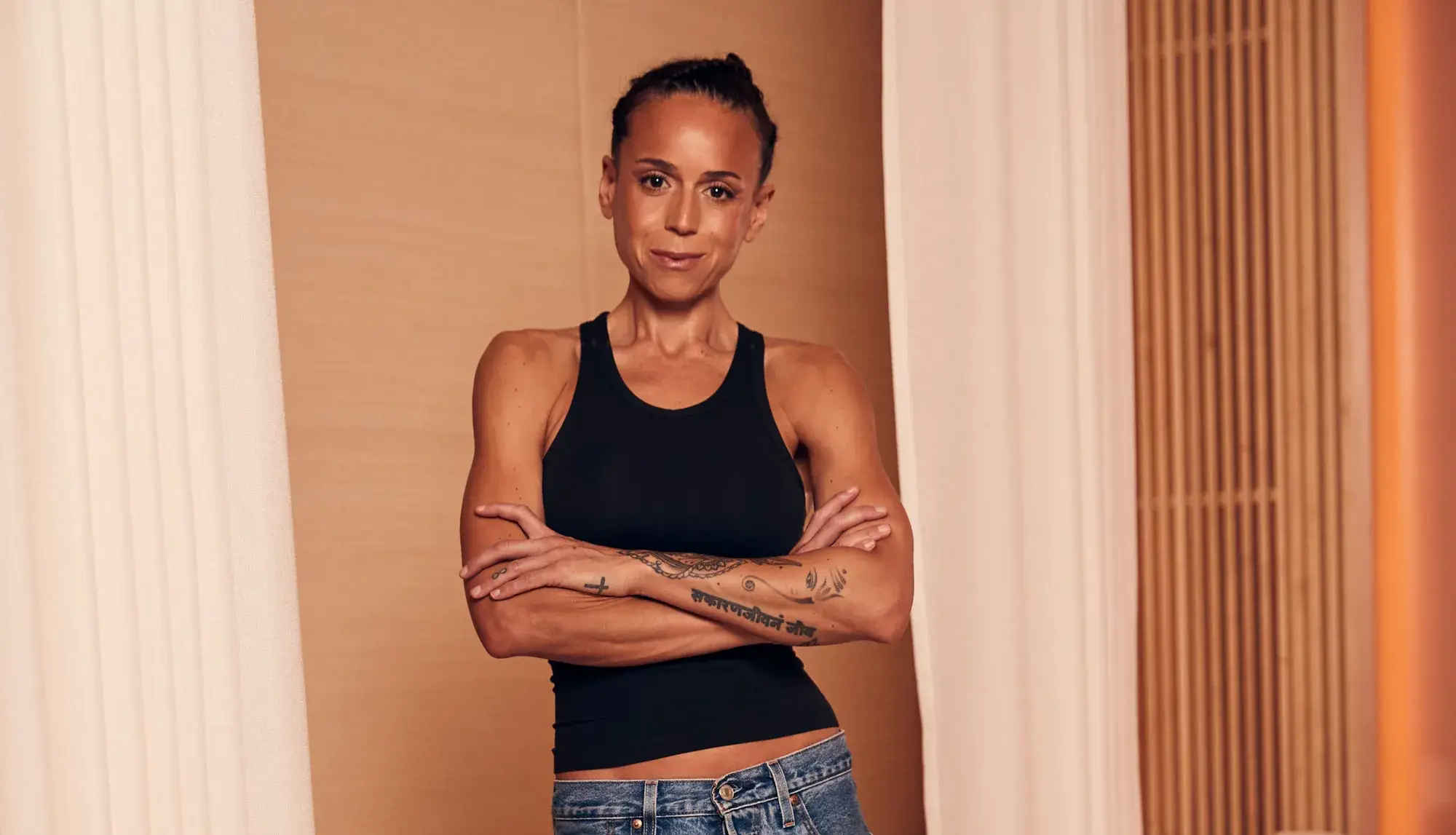 Woman in black tank top standing with arms crossed, showing forearm tattoos against warm neutral interior backdrop.
