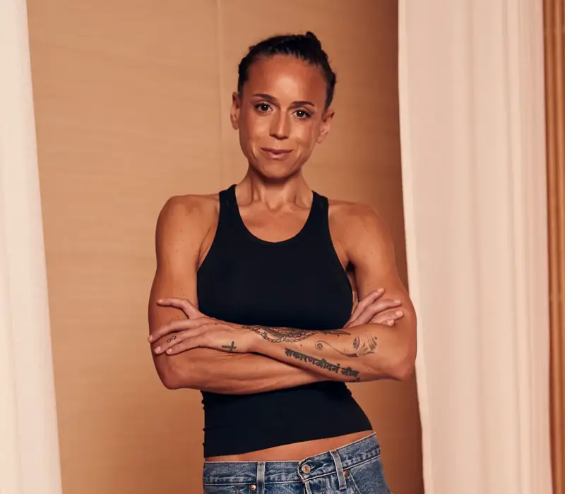 Woman in black tank top standing with arms crossed, showing forearm tattoos against warm neutral interior backdrop.