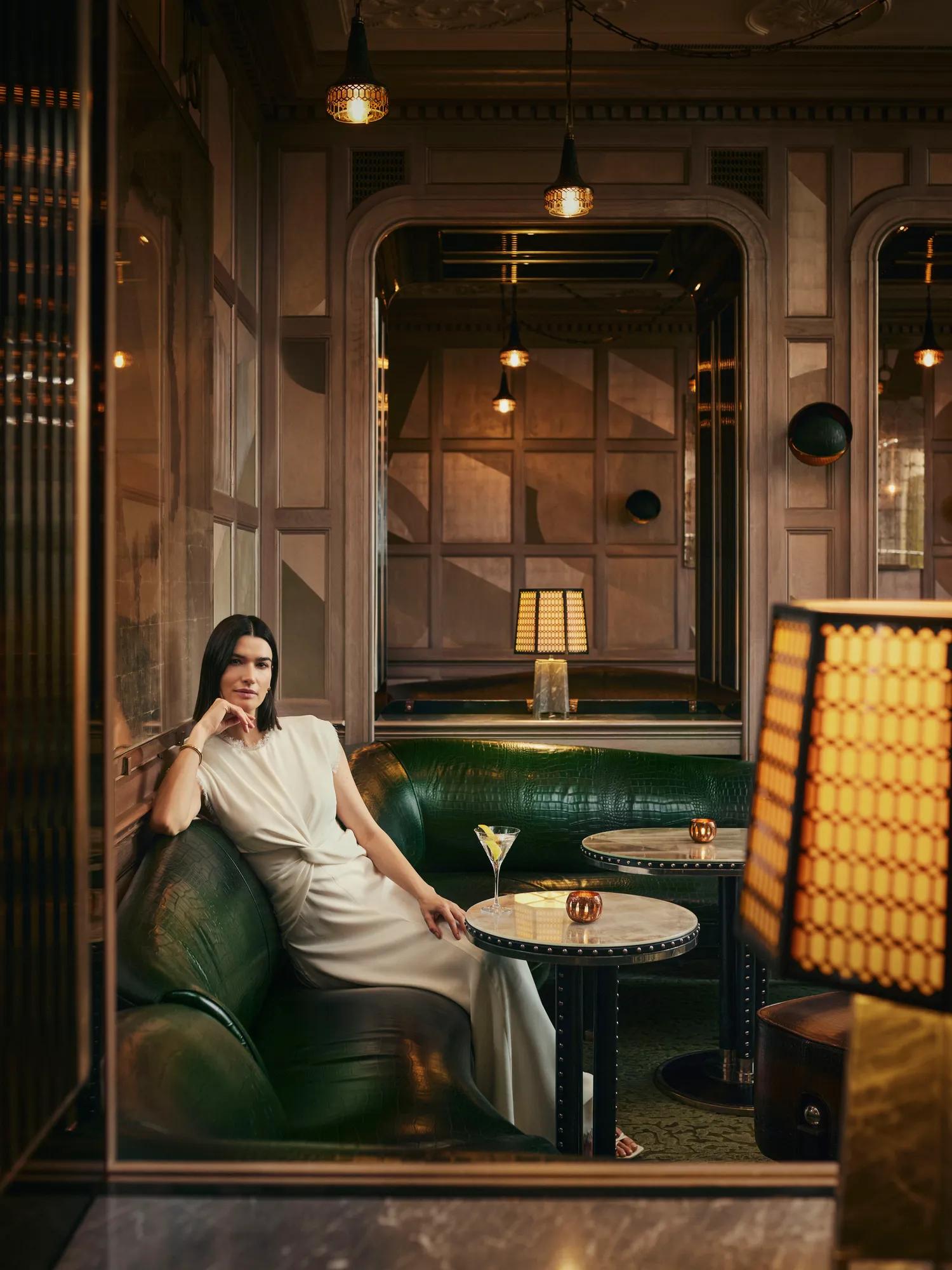 Guest seated on green leather banquette in softly lit Art Deco lounge with cocktail tables and warm ambient lighting.