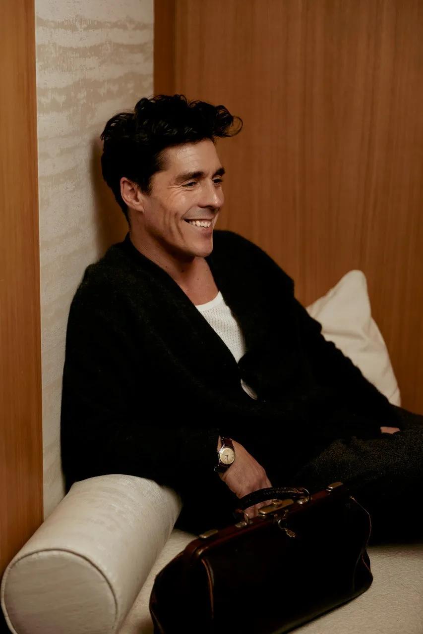 Smiling guest relaxing on a cushioned bench, wearing a dark jacket, with a leather bag resting beside him.