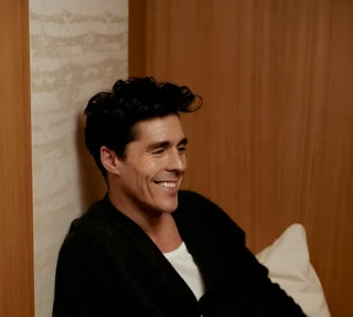 Smiling guest relaxing on a cushioned bench, wearing a dark jacket, with a leather bag resting beside him.