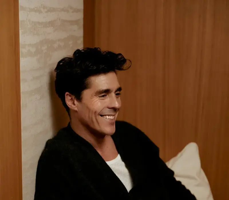 Smiling guest relaxing on a cushioned bench, wearing a dark jacket, with a leather bag resting beside him.