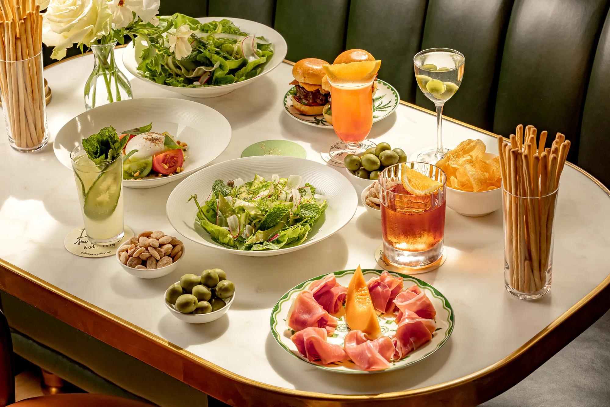Marble table set with salads, charcuterie, olives, breadsticks, and cocktails in a refined restaurant setting.