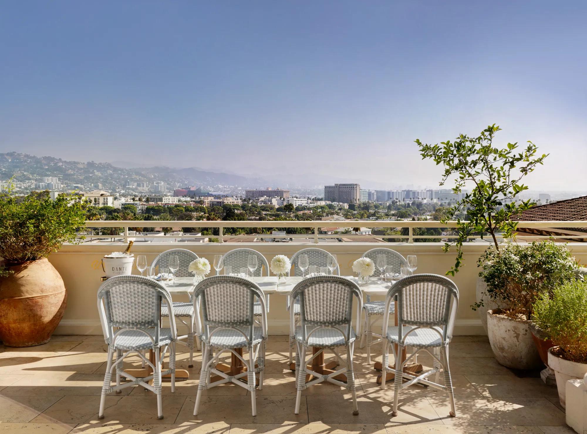 Daytime Dante terrace dining: long table with pale bistro chairs and white florals facing wide city and mountain views.