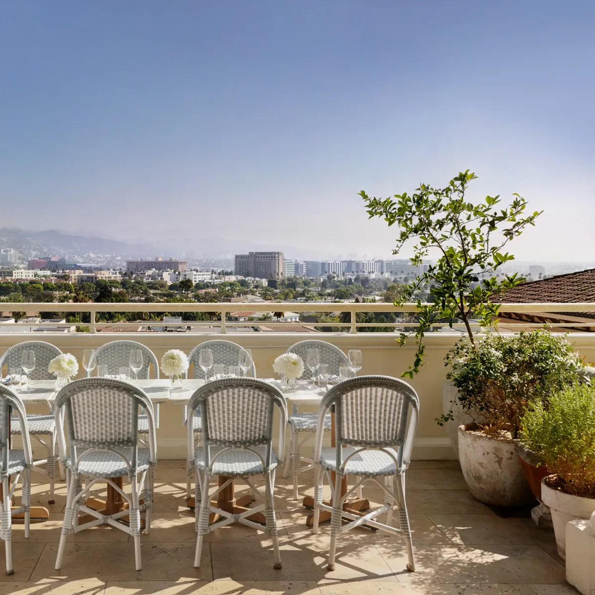 Daytime Dante terrace dining: long table with pale bistro chairs and white florals facing wide city and mountain views.