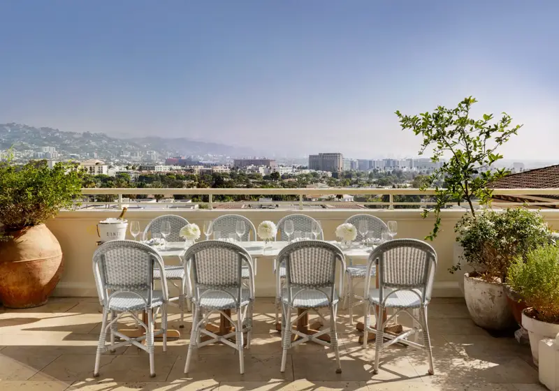 Daytime Dante terrace dining: long table with pale bistro chairs and white florals facing wide city and mountain views.