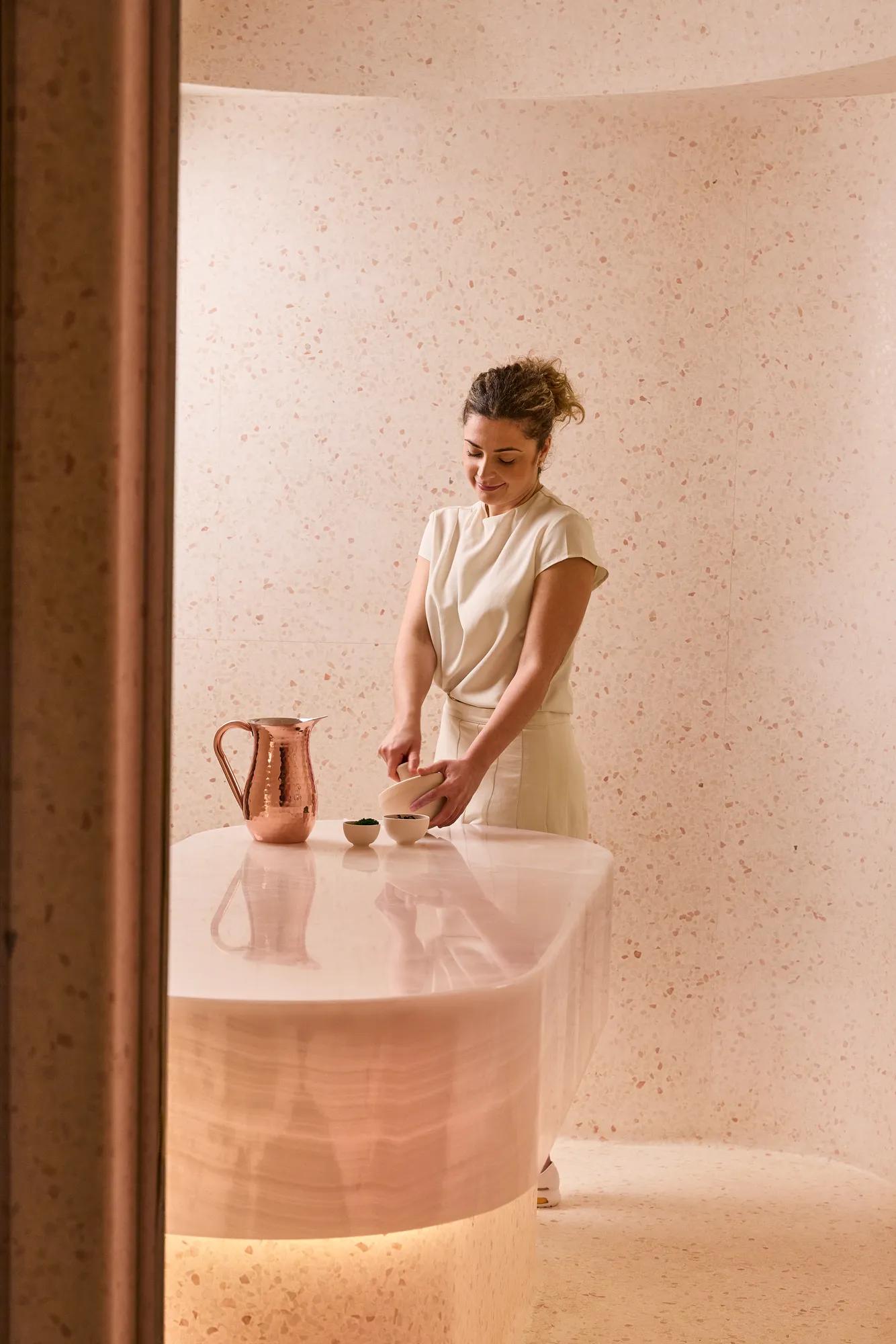 Spa therapist preparing a treatment at a curved stone counter with copper jug and bowls in a softly lit pink-toned room.