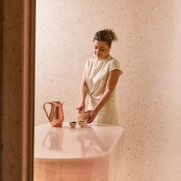 Spa therapist preparing a treatment at a curved stone counter with copper jug and bowls in a softly lit pink-toned room.