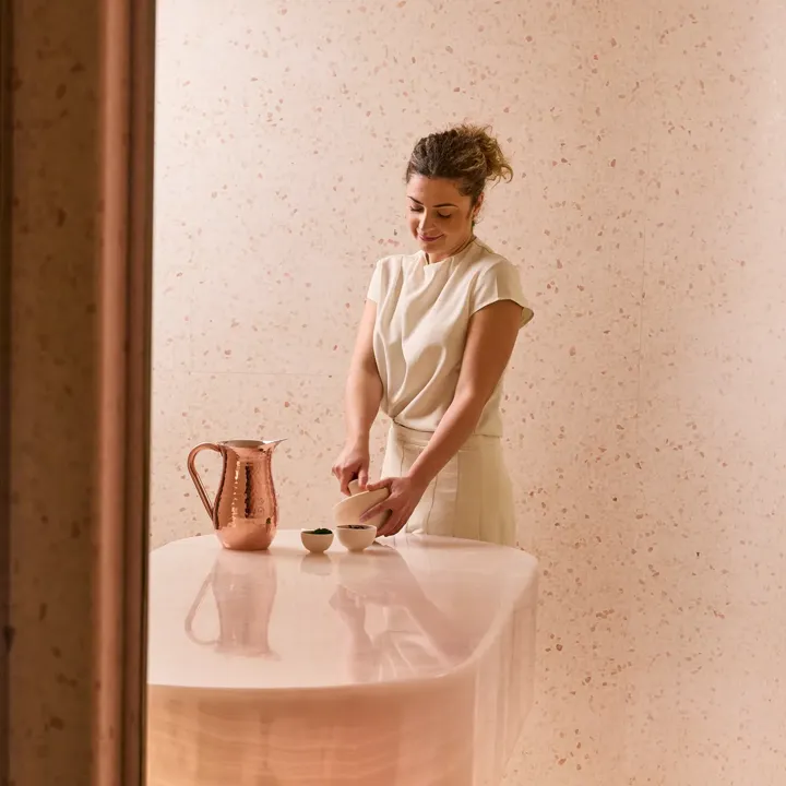 Spa therapist preparing a treatment at a curved stone counter with copper jug and bowls in a softly lit pink-toned room.