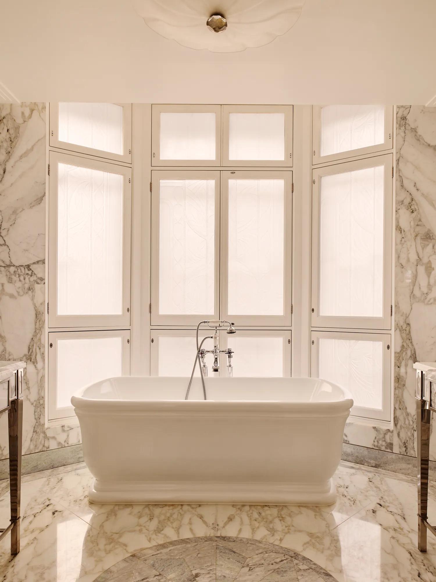 Luxurious marble bathroom with a freestanding white bathtub positioned in front of tall frosted windows, surrounded by soft beige and grey veined marble surfaces.