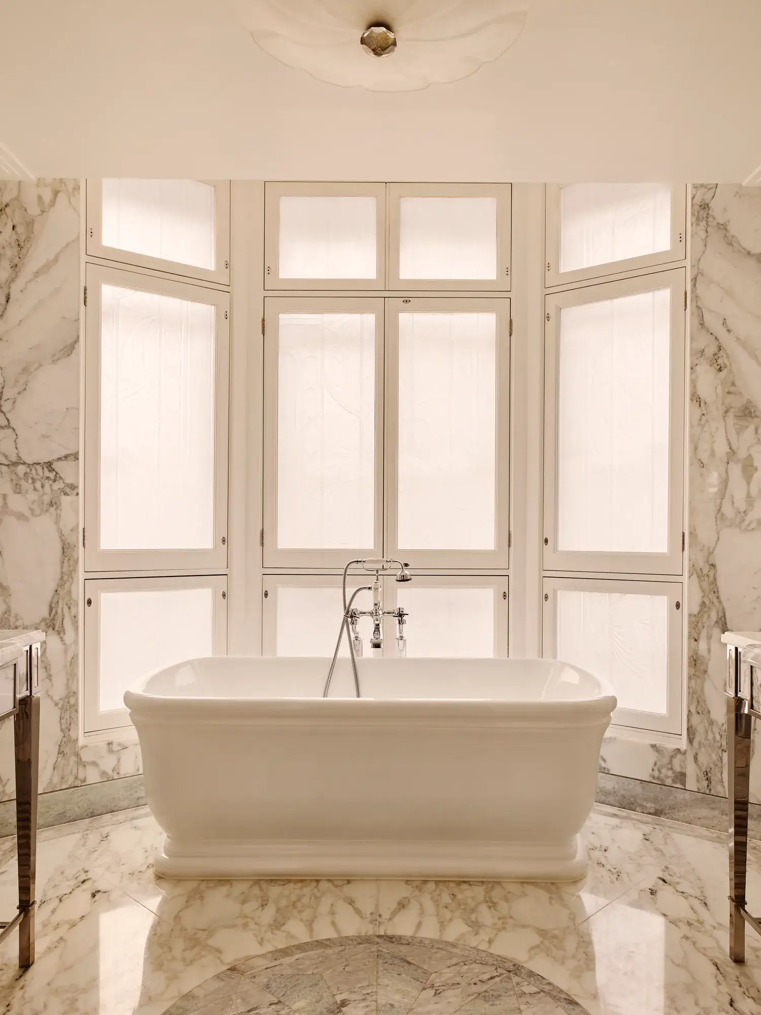 Luxurious marble bathroom with a freestanding white bathtub positioned in front of tall frosted windows, surrounded by soft beige and grey veined marble surfaces.