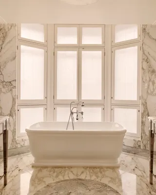 Luxurious marble bathroom with a freestanding white bathtub positioned in front of tall frosted windows, surrounded by soft beige and grey veined marble surfaces.