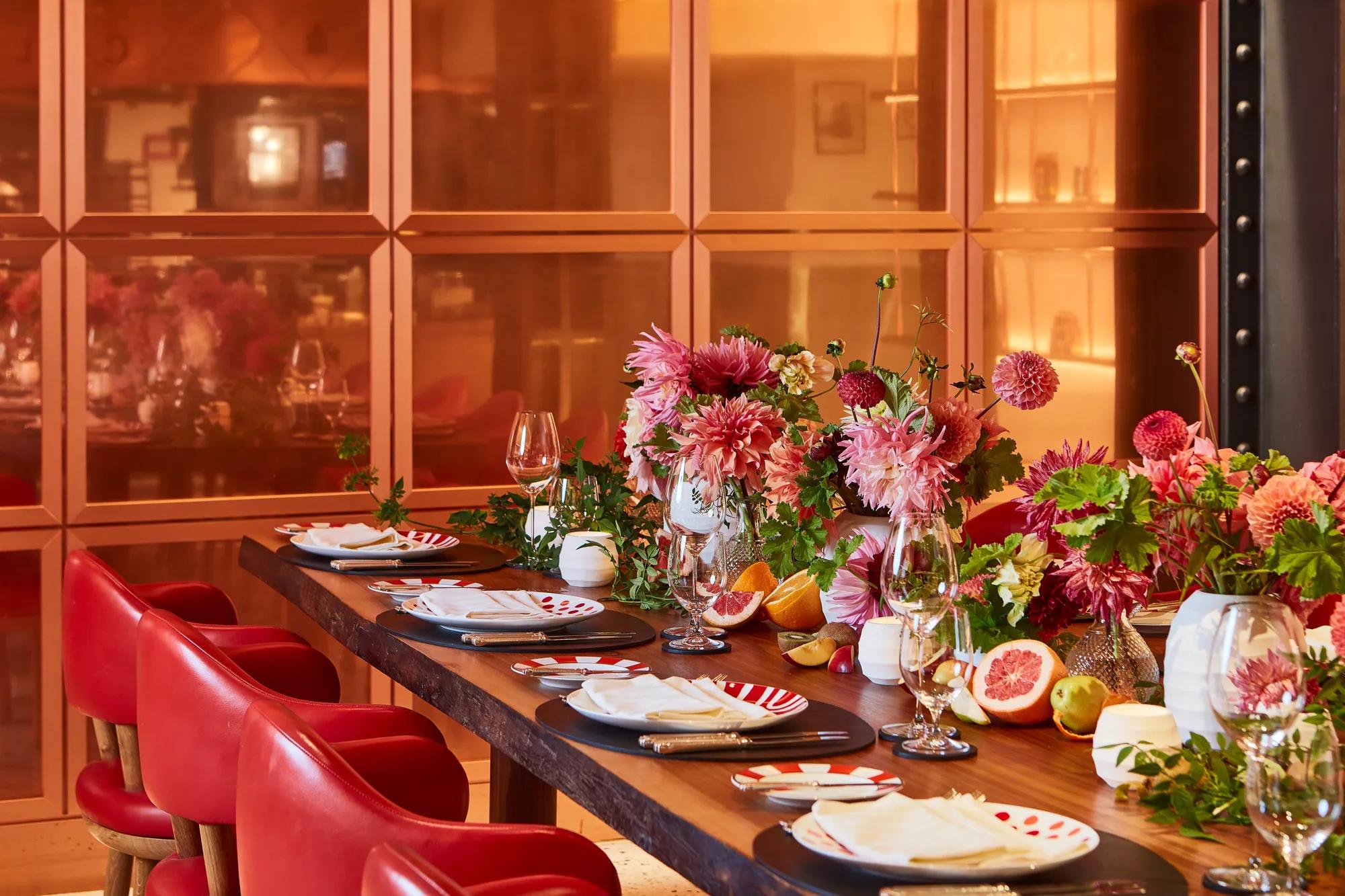 Vibrant dining table set with red chairs, patterned plates, and white napkins, adorned with lush pink and red floral arrangements and fresh fruit. The warm orange-toned backdrop and soft lighting create an inviting, celebratory atmosphere.