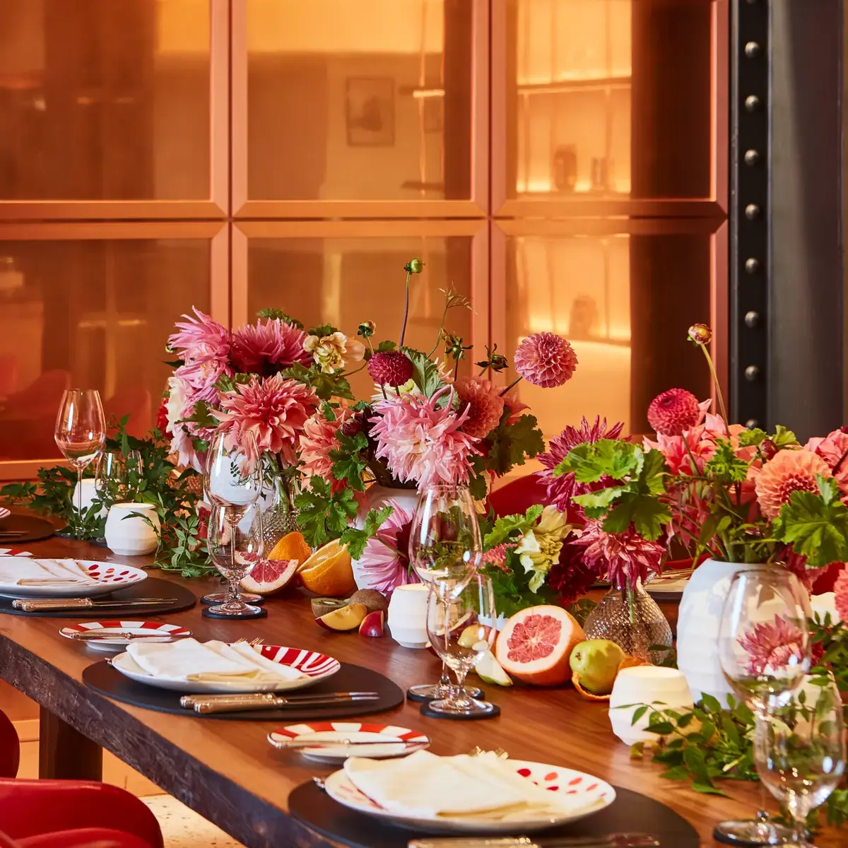 Vibrant dining table set with red chairs, patterned plates, and white napkins, adorned with lush pink and red floral arrangements and fresh fruit. The warm orange-toned backdrop and soft lighting create an inviting, celebratory atmosphere.