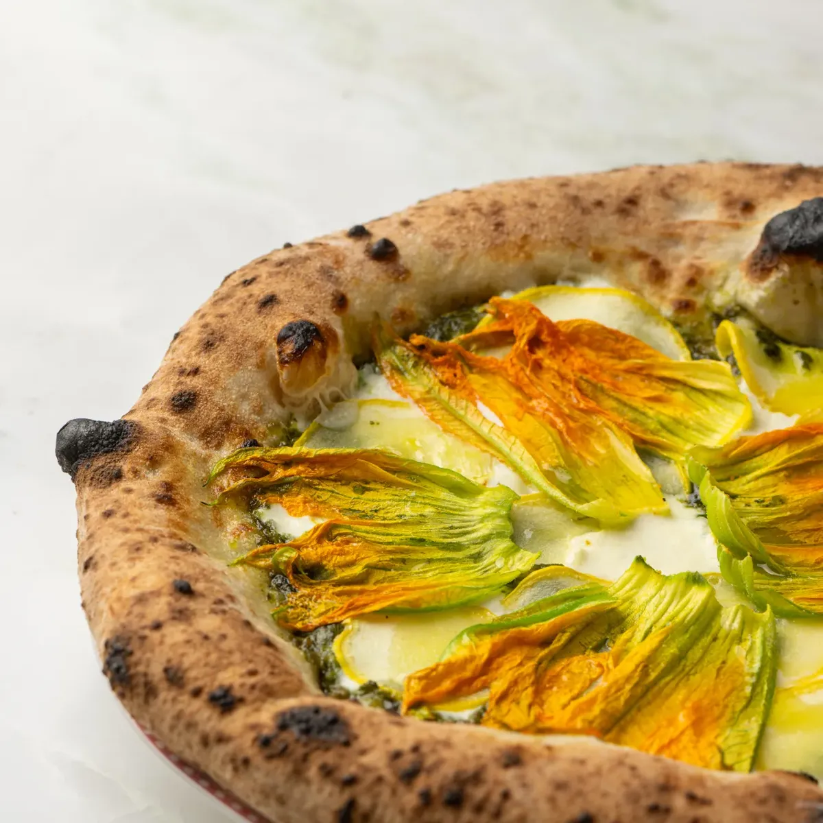 Wood-fired Dante pizza topped with zucchini blossoms and melted cheese on marble surface.