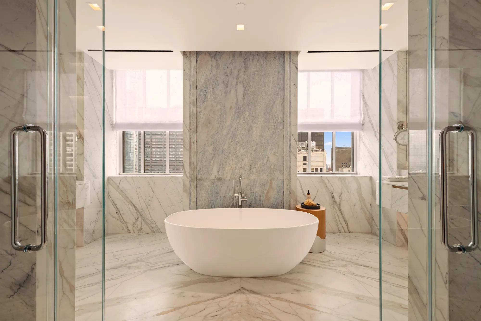 Spacious marble bathroom with freestanding bathtub, twin glass showers and city views framed by large windows.