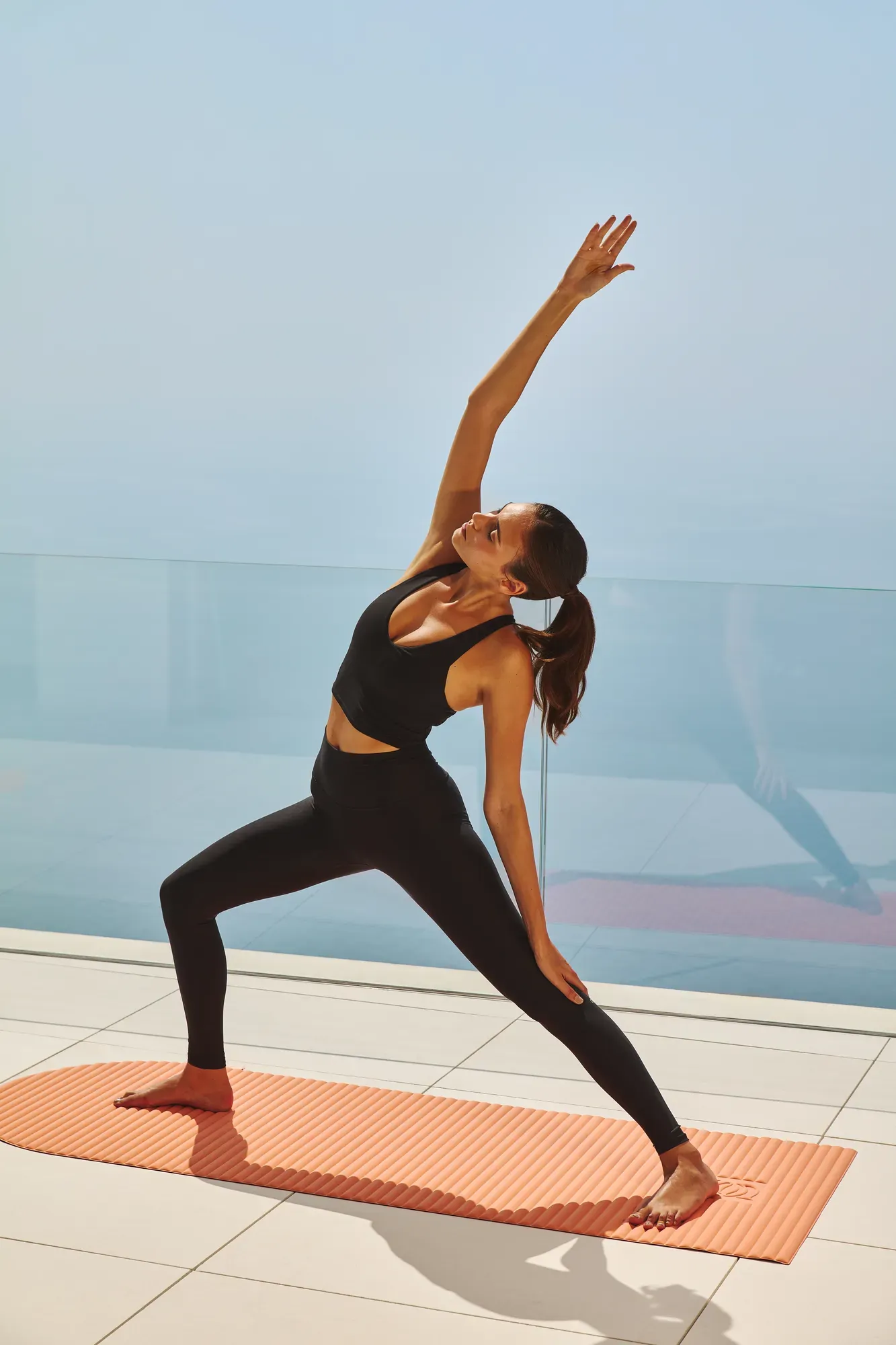Woman practicing yoga on an outdoor terrace with sea views, standing in a side stretch pose on an orange mat.