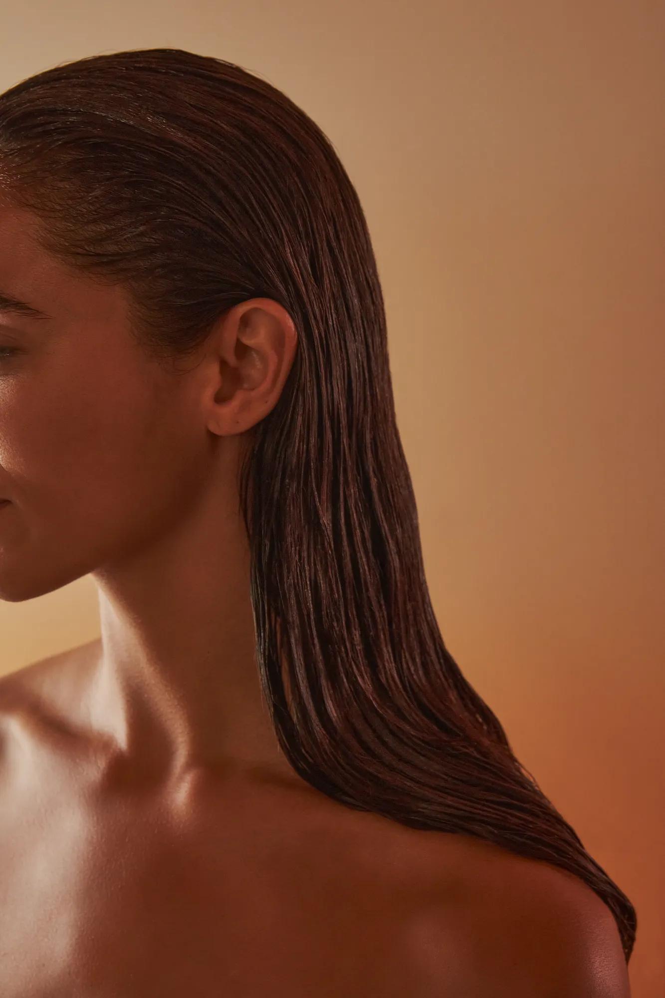 Side profile of woman with slicked-back wet hair and bare shoulders, softly lit against a warm, neutral background.