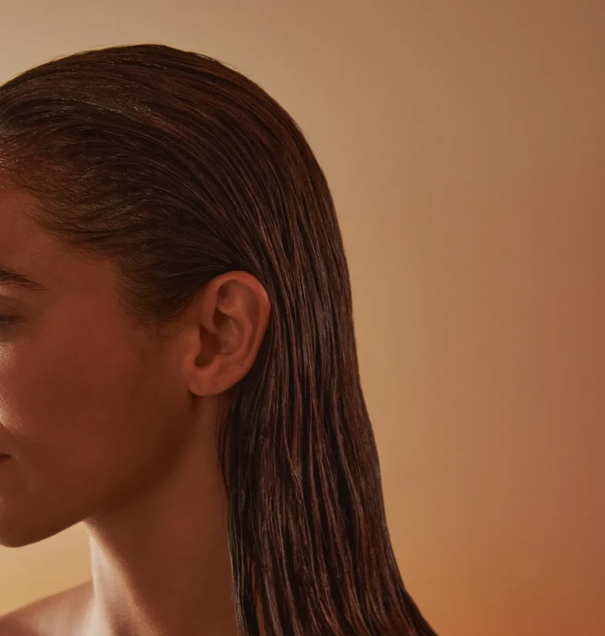 Side profile of woman with slicked-back wet hair and bare shoulders, softly lit against a warm, neutral background.