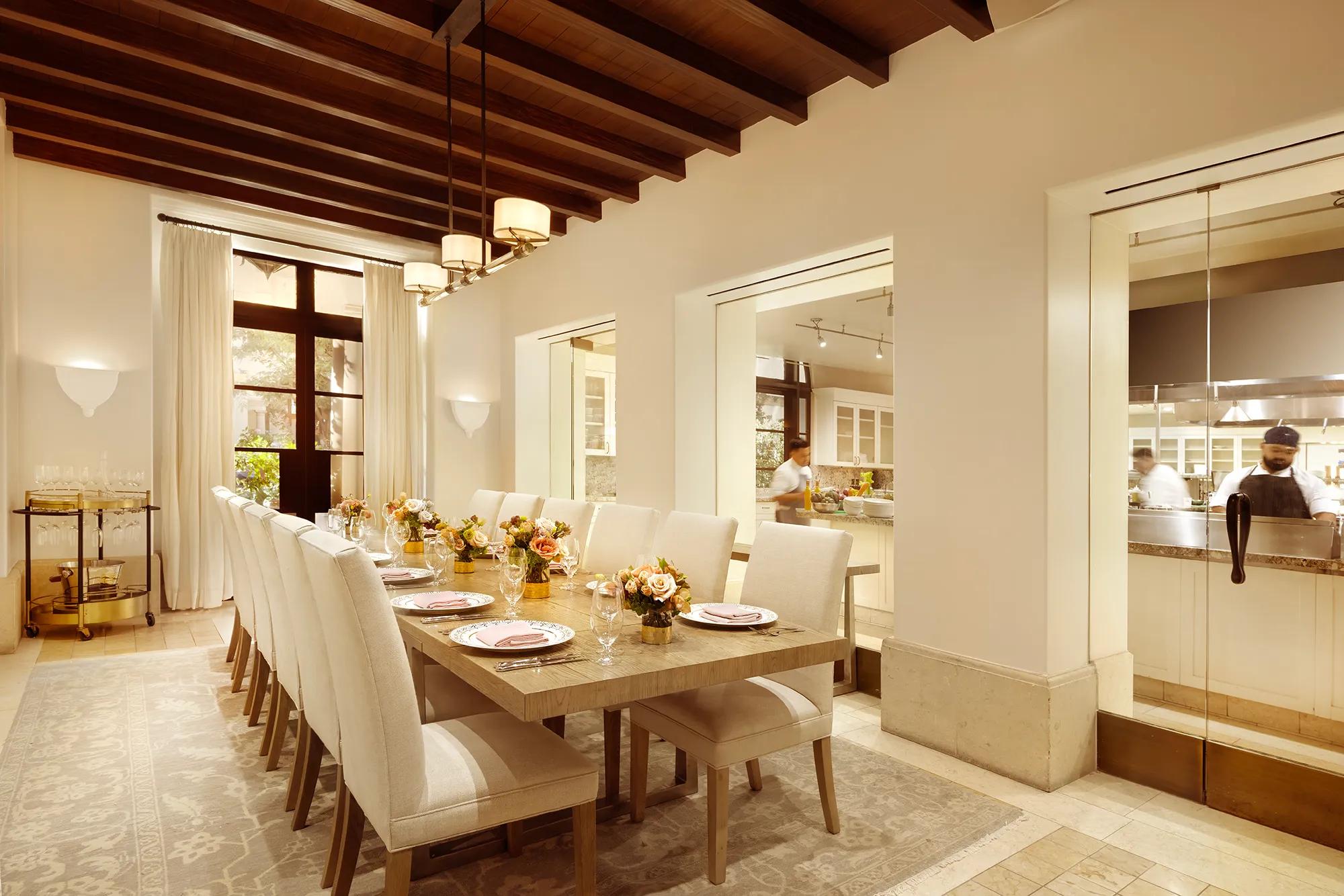 Private dining room at Maybourne Beverly Hills with a long table, upholstered chairs, fresh flower centerpieces, and a view into the open kitchen through glass windows.