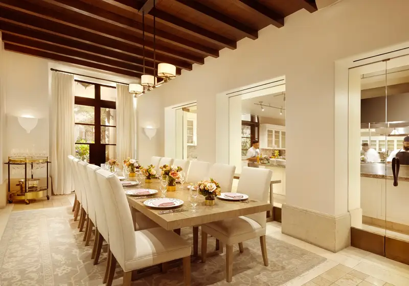 Private dining room at Maybourne Beverly Hills with a long table, upholstered chairs, fresh flower centerpieces, and a view into the open kitchen through glass windows.