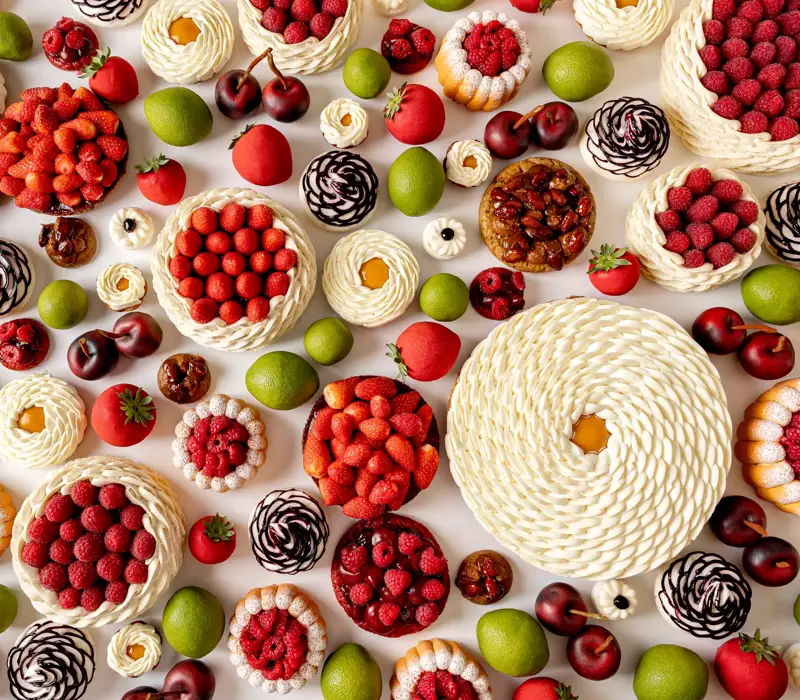 Overhead view of intricate fruit-themed pastries in red, green, and white tones on a white surface.