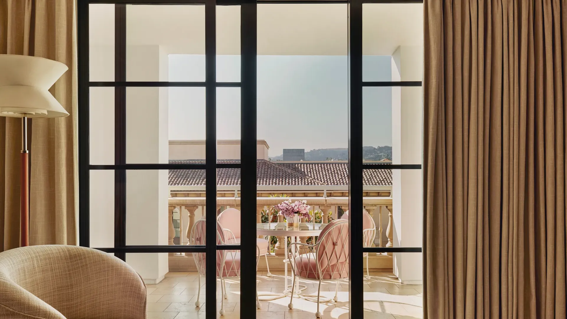 Terrace seen through sliding doors: round table with flowers and four cushioned chairs, with tiled roofs and hills beyond.