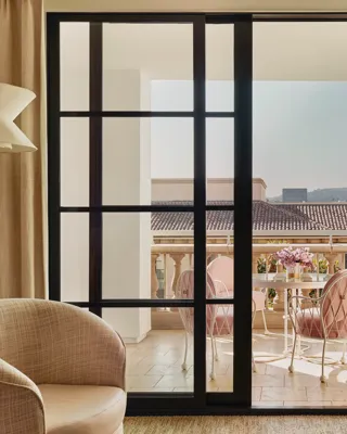 Terrace seen through sliding doors: round table with flowers and four cushioned chairs, with tiled roofs and hills beyond.