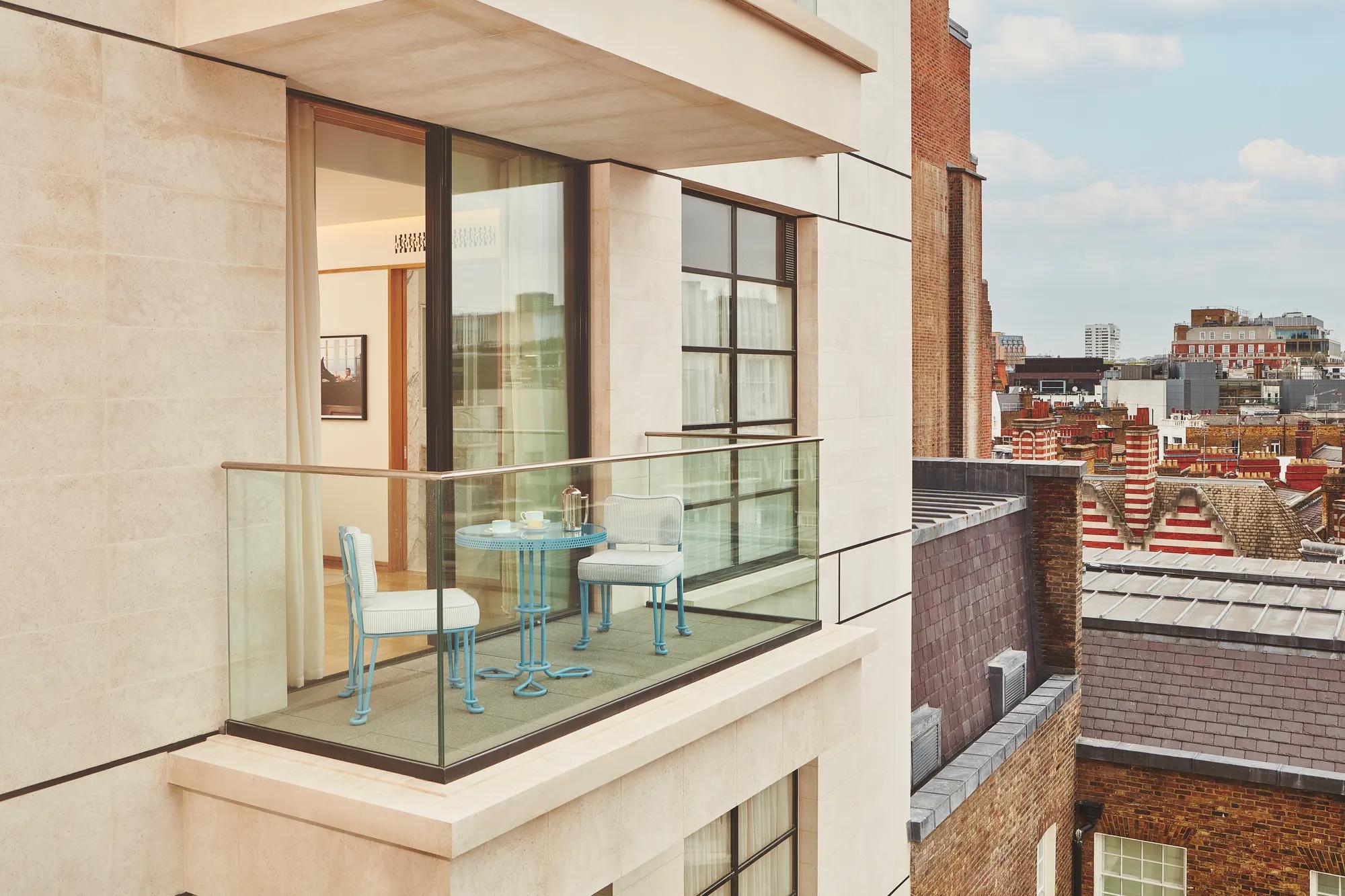 Private balcony with bistro table and chairs, glass balustrade, open doors to bedroom, and city rooftops beyond.
