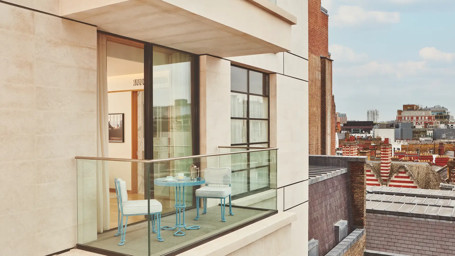 Private balcony with bistro table and chairs, glass balustrade, open doors to bedroom, and city rooftops beyond.