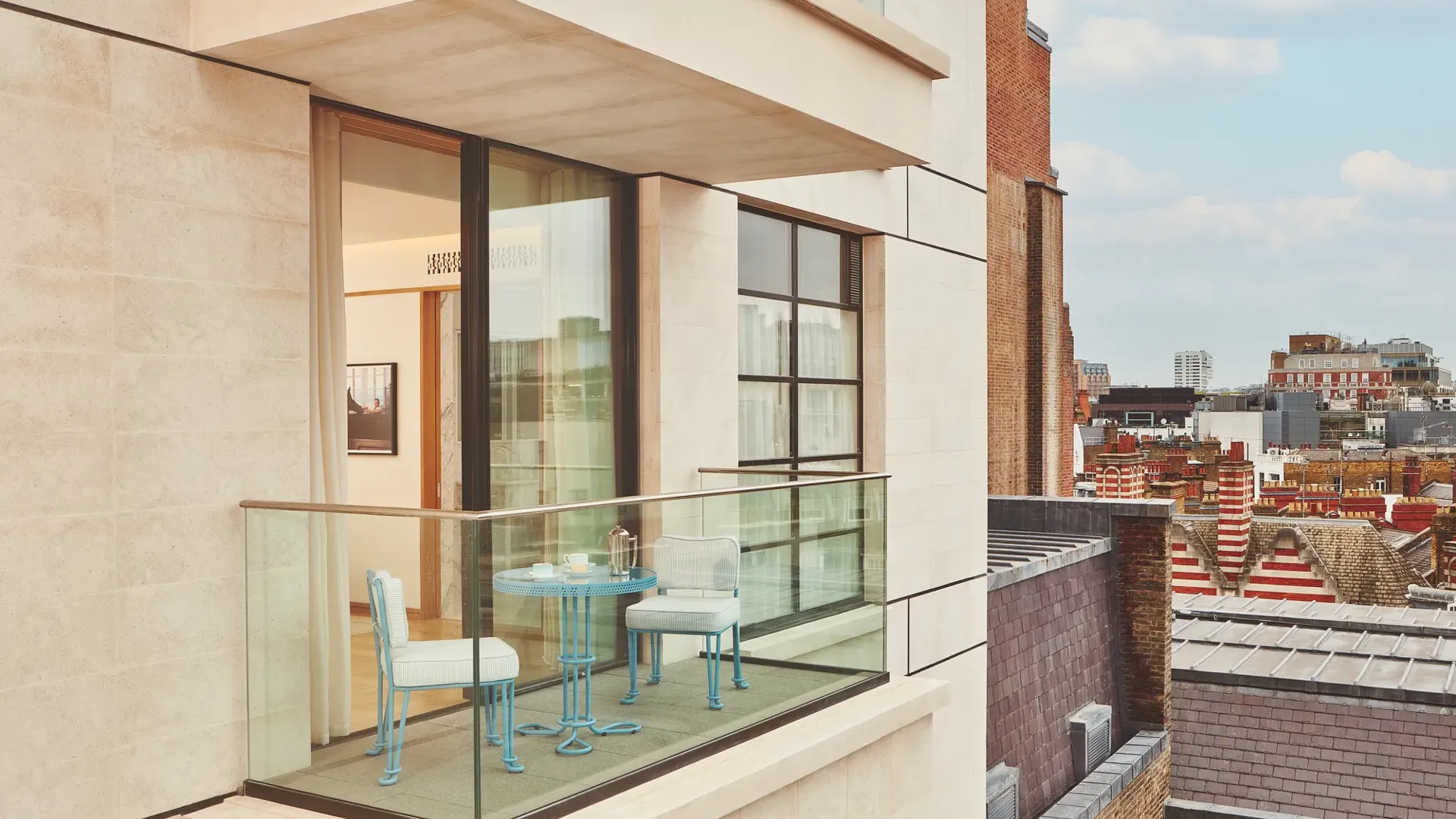Private balcony with bistro table and chairs, glass balustrade, open doors to bedroom, and city rooftops beyond.