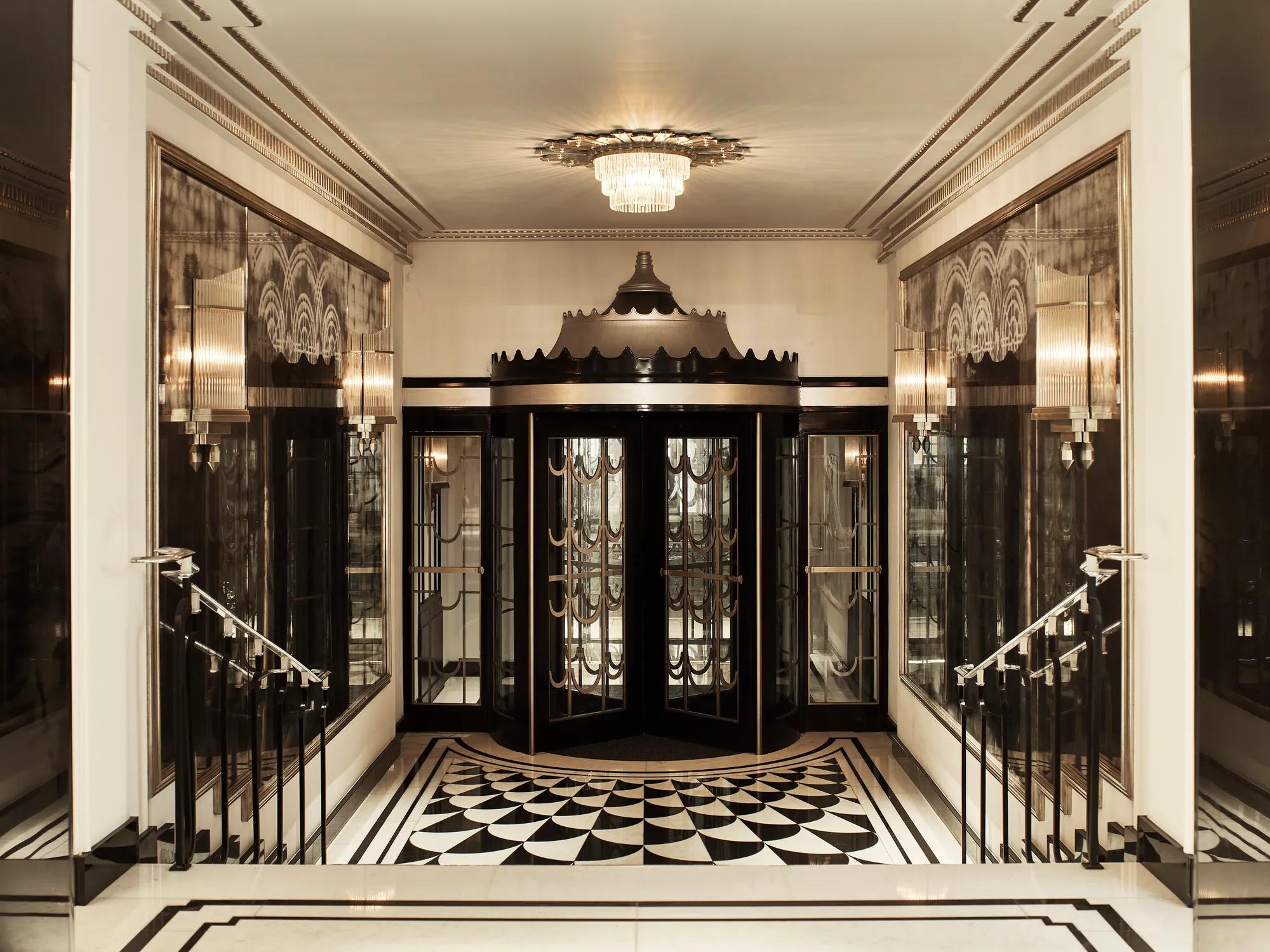 Art Deco revolving door entrance at Claridge’s, featuring black and gold detailing, mirrored walls, geometric black-and-white floor tiles, and a crystal chandelier overhead.