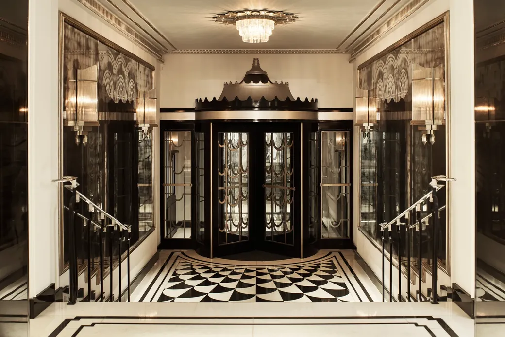 Art Deco revolving door entrance at Claridge’s, featuring black and gold detailing, mirrored walls, geometric black-and-white floor tiles, and a crystal chandelier overhead.