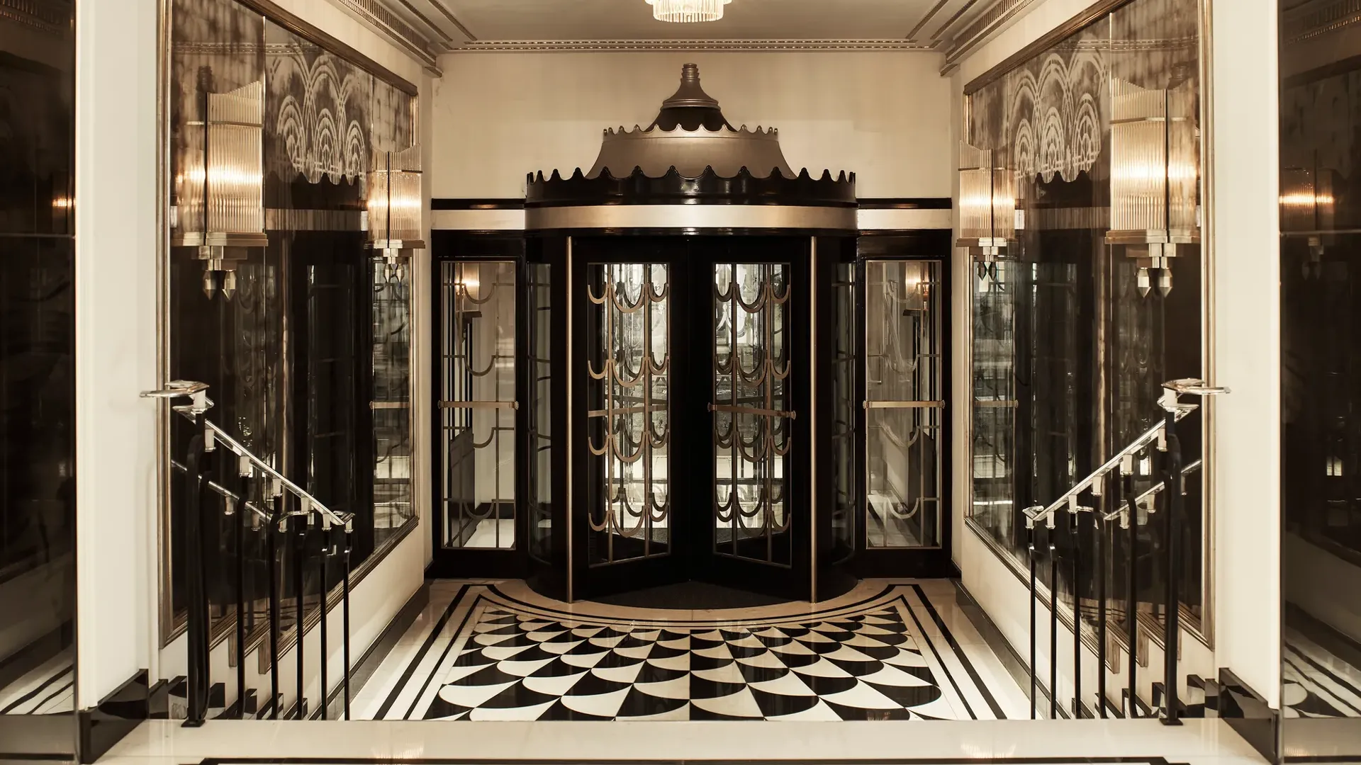 Art Deco revolving door entrance at Claridge’s, featuring black and gold detailing, mirrored walls, geometric black-and-white floor tiles, and a crystal chandelier overhead.