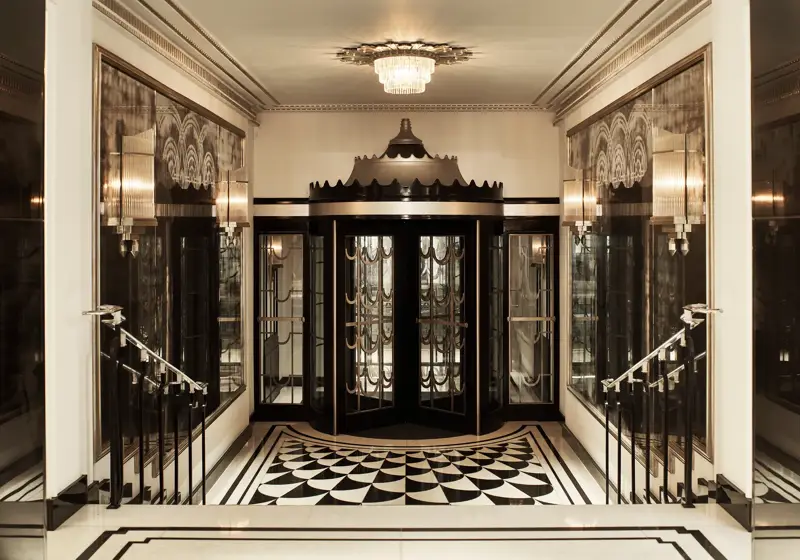Art Deco revolving door entrance at Claridge’s, featuring black and gold detailing, mirrored walls, geometric black-and-white floor tiles, and a crystal chandelier overhead.