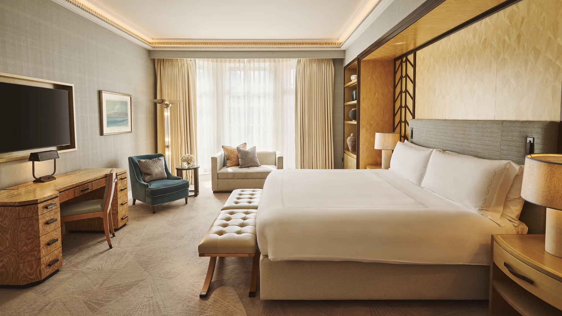 Spacious bedroom with king bed and bench, desk with TV, armchair and sofa, floor-to-ceiling window, and warm lighting.