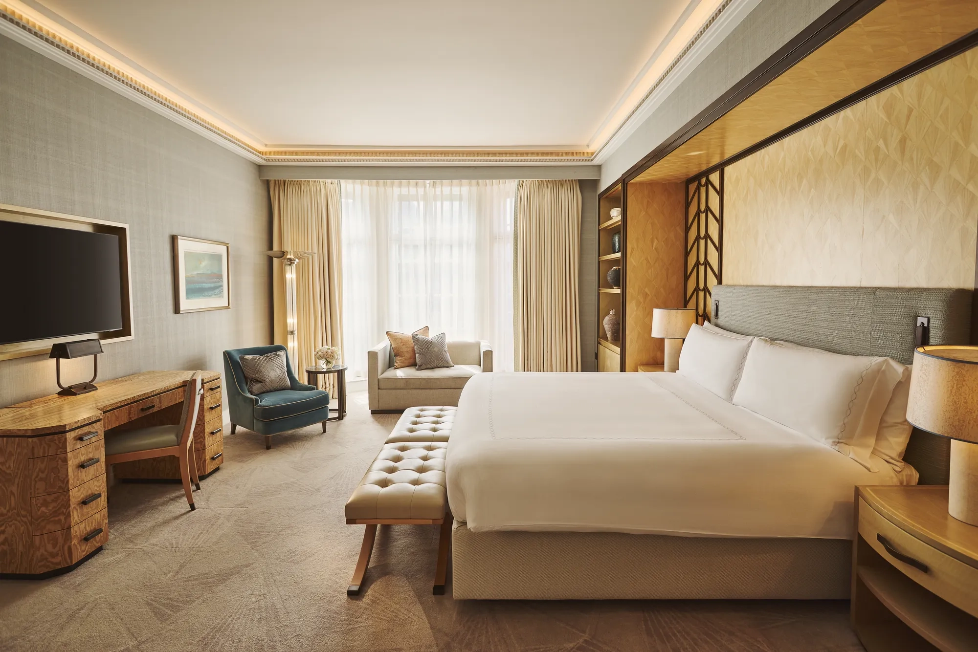 Spacious bedroom with king bed and bench, desk with TV, armchair and sofa, floor-to-ceiling window, and warm lighting.