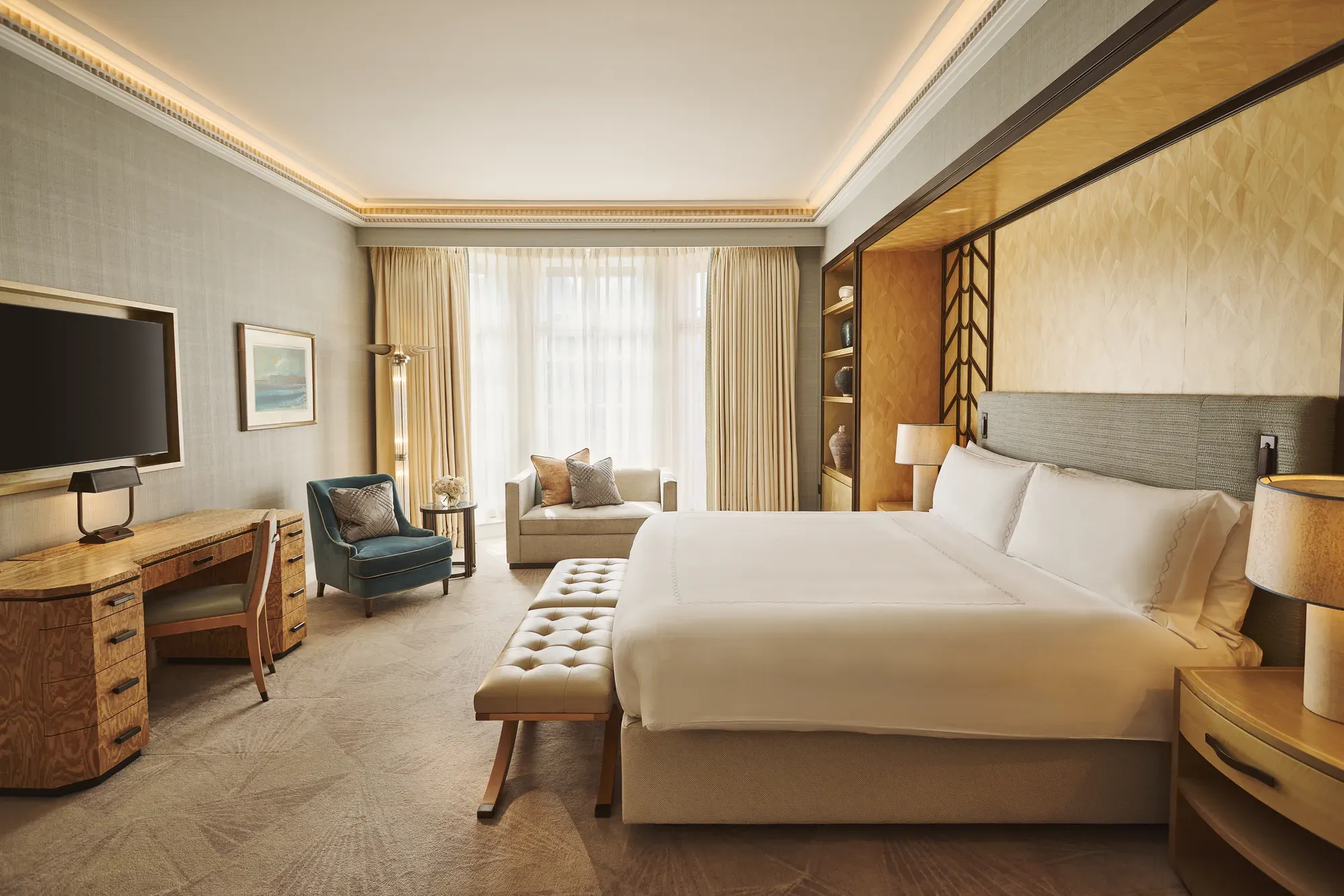 Spacious bedroom with king bed and bench, desk with TV, armchair and sofa, floor-to-ceiling window, and warm lighting.