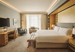 Spacious bedroom with king bed and bench, desk with TV, armchair and sofa, floor-to-ceiling window, and warm lighting.