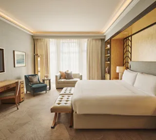 Spacious bedroom with king bed and bench, desk with TV, armchair and sofa, floor-to-ceiling window, and warm lighting.