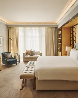 Spacious bedroom with king bed and bench, desk with TV, armchair and sofa, floor-to-ceiling window, and warm lighting.