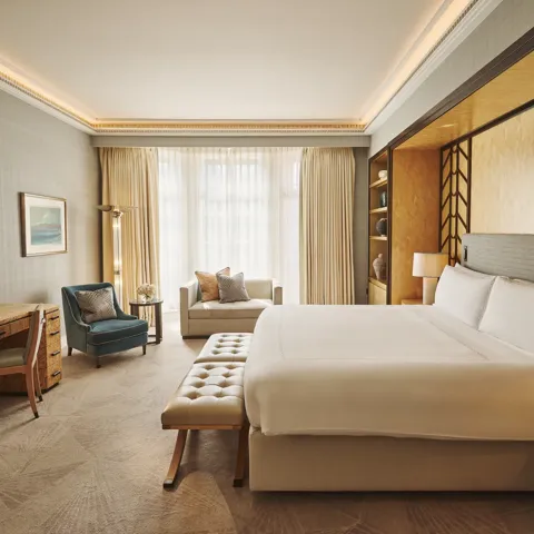 Spacious bedroom with king bed and bench, desk with TV, armchair and sofa, floor-to-ceiling window, and warm lighting.