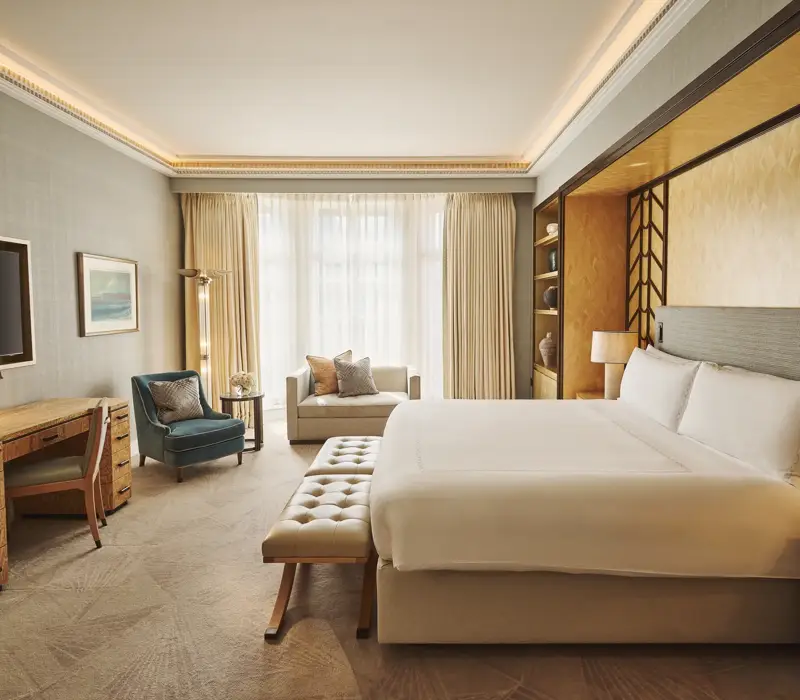 Spacious bedroom with king bed and bench, desk with TV, armchair and sofa, floor-to-ceiling window, and warm lighting.