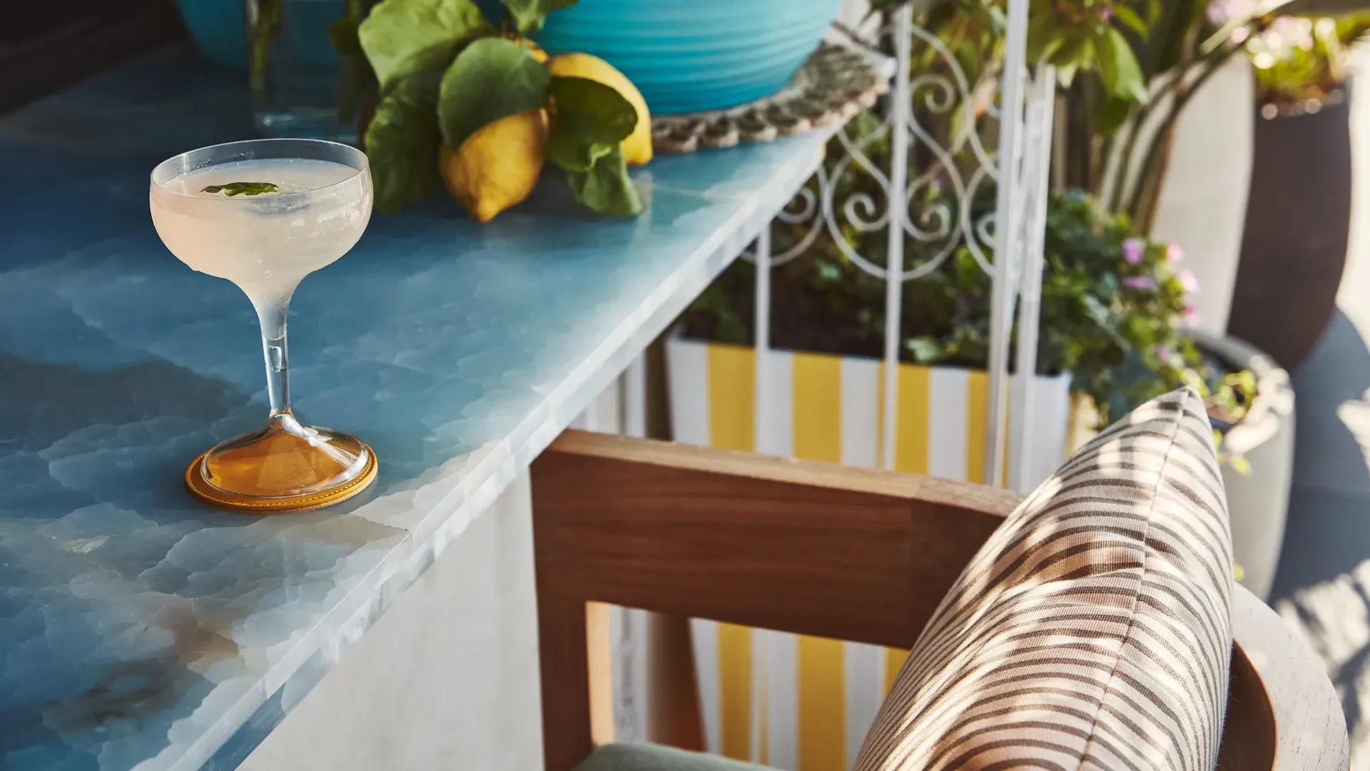 Pale cocktail in a coupe glass on a blue marble bar beside lemons and lush greenery, with a striped cushion chair in sunlight.