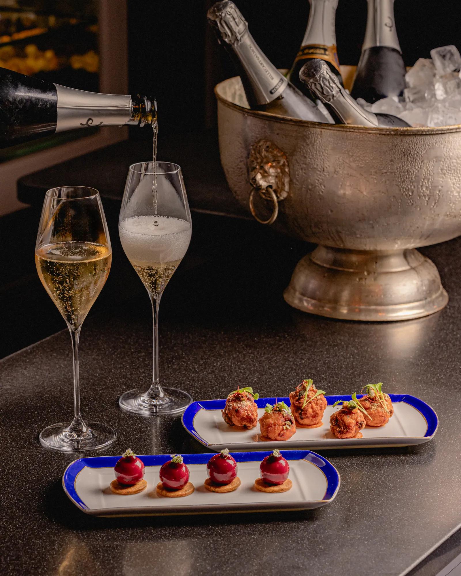 Champagne being poured into flutes beside an ice bucket, with two plates of bite-sized canapés on a bar counter.