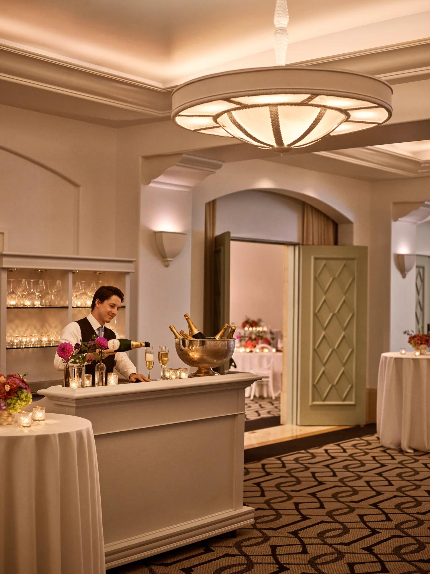 Ballroom foyer bar with bartender pouring champagne, votive candles, and floral décor in warm ambient lighting.
